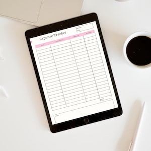 Könnte beinhalten: Ein schwarzes Tablet mit einem "Expense Tracker"-Formular mit rosa Überschriften und einem Raster für die Eingabe von Finanzdaten. Ein Laptop, Kaffee, Stift und Ornament befinden sich auf einer weißen Oberfläche.