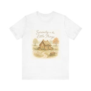 Könnte beinhalten: Weißes T-Shirt mit einer Aquarellillustration eines Cottage in einem Feld und dem Text "Serenity in the Little Things". Das Bild zeigt einen gewundenen Pfad, Bäume und einen kleinen Wagen.