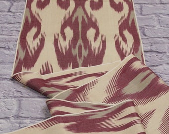 Può includere: Un primo piano di un tessuto beige e bordeaux con un motivo ikat tradizionale. Il tessuto è piegato e drappeggiato, mostrando il design intricato.