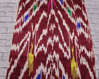 May include: A close-up of a burgundy fabric with a white and yellow ikat pattern. The fabric has a subtle sheen and a soft texture.