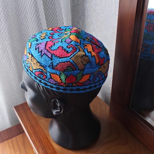 以下が含まれることがあります： 青、赤、オレンジ、紫の色合いの鮮やかな刺繍スカルキャップ。帽子は詳細な幾何学模様が特徴で、黒いマネキンの頭に展示されています。このキャップは伝統的なスタイルで、おそらく宗教的または文化的な着用を目的としています。
