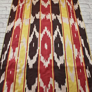 Può includere: Tessuto a righe con un motivo ripetuto di rosso, giallo, bianco e nero. Il tessuto ha una trama tessuta e un design ikat tradizionale.