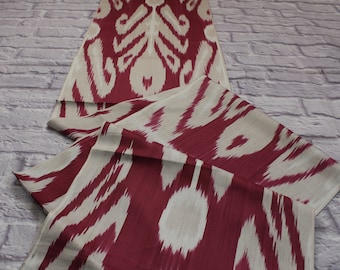 May include: Red and white ikat fabric with a traditional pattern. The fabric is folded and draped on a white brick surface.