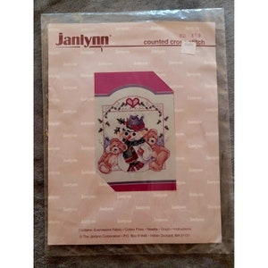 May include: A counted cross stitch pattern featuring a snowman with two teddy bears. The pattern is on a pink background with the text "Janlynn" and "counted cross stitch" printed on the top. The pattern includes instructions for making the cross stitch.