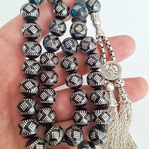 Puede incluir: Un collar de cuentas de oración negras con detalles plateados y un borla plateada. Las cuentas son redondas y tienen un patrón geométrico.