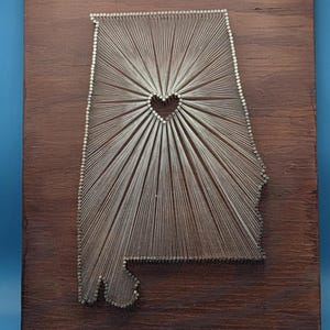 Könnte beinhalten: Wandkunst aus Holz mit der Form von Alabama, hergestellt aus Nägeln und Faden. Das Design zeigt ein Herz in der Mitte mit strahlenförmigen Linien. Die Holzbasis hat eine dunkelbraune Oberfläche und misst etwa 30 cm x 30 cm.