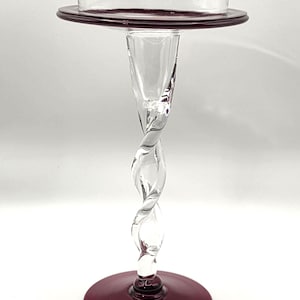 May include: A clear glass candlestick with a twisted stem and a circular base and top in a deep burgundy color. The candlestick has a classic design, suitable for holding a candle.