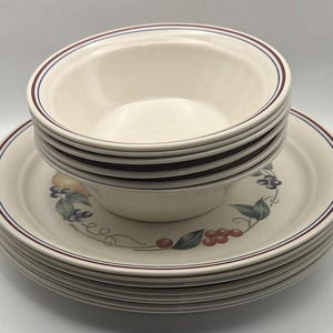 May include: A set of off-white ceramic bowls and plates. The bowls and plates have a maroon and navy blue trim. The plates feature a fruit and leaf design. The bowls are stacked on top of the plates.