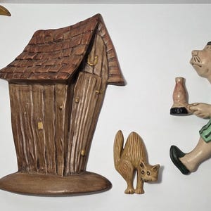 May include: A whimsical wall art set featuring a crescent moon, a howling dog, a small outhouse, a scared cat, and a figure in striped pajamas carrying a bottle. The figures are painted in warm, earthy tones.