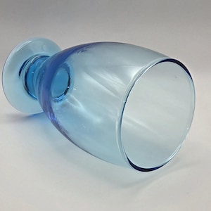 May include: A light blue glass vase with a wide, rounded body and a flared base. The vase has a clear, transparent appearance, with a darker blue rim and base. The vase is lying on its side, showcasing its shape and design.