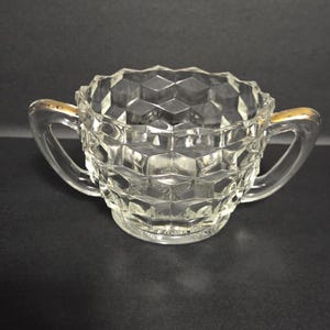 Puede incluir: Azucarero de cristal transparente con un patrón geométrico de diamantes. El cuenco tiene dos asas curvas y un borde dorado. El azucarero está sobre una superficie oscura.