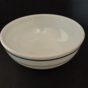 May include: A white ceramic bowl with a simple design. The bowl has a rounded shape and is encircled by two thin, parallel blue lines near the rim. The bowl is set against a dark background.