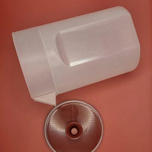 May include: A clear, rectangular container with a handle and a brown, circular lid. The lid has a central, raised knob. The container and lid are set against a solid, reddish-brown background. The container appears to be made of plastic.