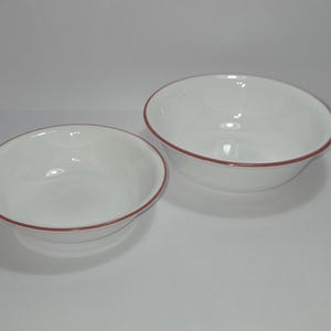 May include: Two white ceramic bowls with a red rim. The bowls are different sizes and are set against a white background. The bowls have a simple, clean design, suitable for serving food.