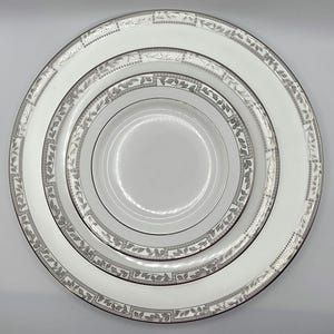 May include: A set of three white ceramic plates, nested together. Each plate has a silver border with a repeating leaf and geometric design. The plates vary in size, with the smallest in the centre.
