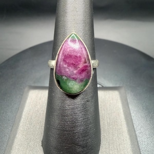 May include: A silver ring featuring a large, teardrop-shaped gemstone. The stone displays a vibrant mix of colors, with a deep magenta center and green edges. The ring is displayed on a black jewelry stand, showcasing its elegant design.