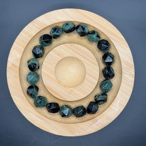 May include: A beaded bracelet featuring faceted, dark green and black gemstones. The bracelet is displayed on a round, wooden, tiered tray with a natural finish. The gemstones have a geometric cut, reflecting light. The background is a dark gray.