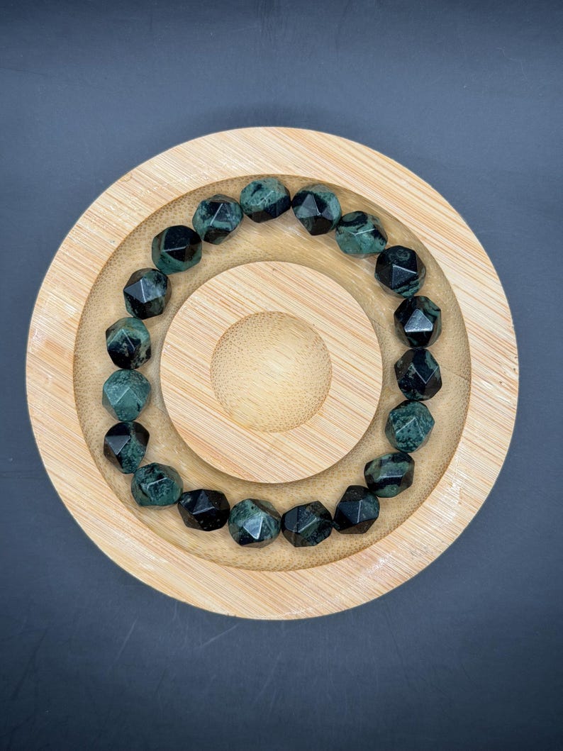 May include: A beaded bracelet featuring faceted, dark green and black gemstones. The bracelet is displayed on a round, wooden, tiered tray with a natural finish. The gemstones have a geometric cut, reflecting light. The background is a dark gray.
