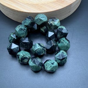 May include: A collection of faceted, dark green gemstone beads. The beads have a geometric shape and a polished surface, with variations in color from light to dark green. The beads are arranged in a circular shape, suggesting they are part of a bracelet or necklace.