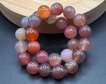 May include: A bracelet made of round, polished beads in shades of orange, pink, and gray. The beads are strung together, forming a circular shape. The bracelet is displayed on a wooden stand against a dark background.
