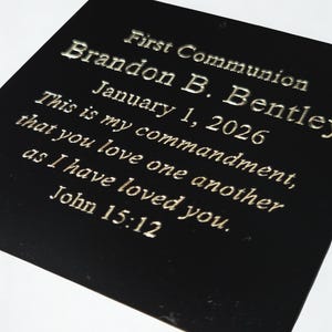 Puede incluir: Placa negra con letras plateadas. El texto dice "Primera Comuni&oacute;n, Brandon B. Bentley, 1 de enero de 2026." Tambi&eacute;n incluye un vers&iacute;culo b&iacute;blico de Juan 15:12. La placa es rectangular y brillante.