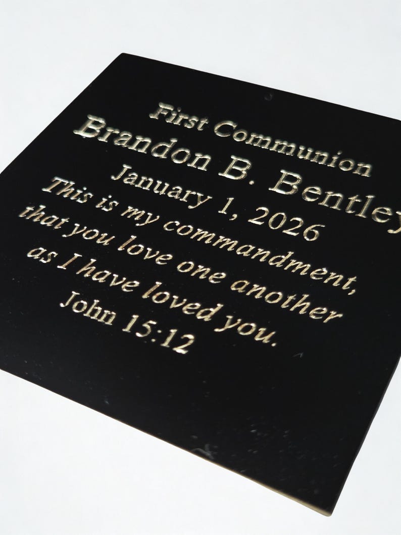 Puede incluir: Placa negra con letras plateadas. El texto dice "Primera Comuni&oacute;n, Brandon B. Bentley, 1 de enero de 2026." Tambi&eacute;n incluye un vers&iacute;culo b&iacute;blico de Juan 15:12. La placa es rectangular y brillante.