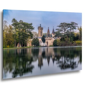 May include: A stone castle with a tall tower and pointed roofs sits on the edge of a calm lake. Tall green trees surround the structure, and the entire scene is reflected in the still water under a cloudy blue sky during the daytime.