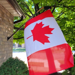 Könnte beinhalten: Eine kanadische Flagge ist an einem Hockeyschläger befestigt und hängt an einem Haus. Die Flagge ist rot und weiß mit einem großen roten Ahornblatt in der Mitte. Die Flagge weht im Wind.