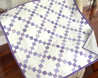 May include: A square quilt featuring a white and purple patchwork pattern with a chain-link design. The quilt is bordered with a solid purple fabric binding and displays a floral patterned fabric on the underside corner, with intricate swirl quilting.