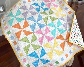 May include: A colorful handmade quilt with a pinwheel design. The quilt features a white background with vibrant triangles in shades of pink, orange, yellow, green, blue, and purple. The border is a yellow binding with colorful squares.