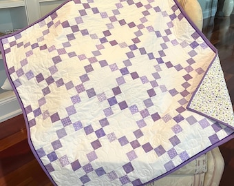 May include: A handmade quilt with a white background and a pattern of purple squares. The squares are arranged in a grid and feature various designs. The quilt is trimmed with a solid purple border and has a floral patterned backing.