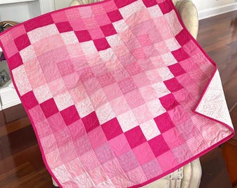 May include: A square patchwork quilt featuring a pixelated heart design in various shades of pink, ranging from light blush to deep magenta. The quilt is constructed from small fabric squares with diverse textures and patterns, finished with a bright pink border.