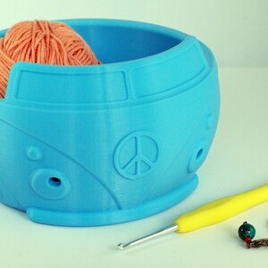 May include: A blue ceramic yarn bowl shaped like a vintage Volkswagen bus with a peace sign on the front. The bowl has holes for yarn to feed through. A ball of orange yarn is in the bowl. A yellow crochet hook and two earrings are on a white surface.