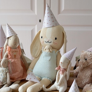 May include: A group of plush toys wearing party hats. The toys include a bunny rabbit, a teddy bear, and a doll. The bunny rabbit is wearing a blue and white striped overall and a pink party hat. The teddy bear is wearing a pink party hat. The doll is wearing a pink and white dress and a pink party hat.