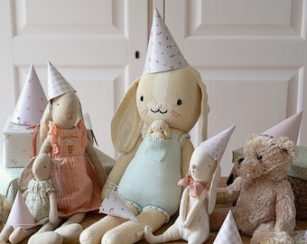 May include: A group of plush toys wearing party hats. The toys include a bunny rabbit, a teddy bear, and a doll. The bunny rabbit is wearing a blue and white striped overall and a pink party hat. The teddy bear is wearing a pink party hat. The doll is wearing a pink and white dress and a pink party hat.