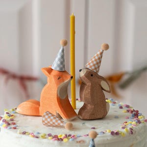 May include: A white frosted cake with colourful sprinkles, topped with a wooden fox and rabbit wearing party hats. The fox is orange and the rabbit is brown.
