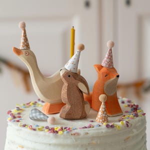 May include: A white cake with colorful sprinkles is decorated with wooden animal figurines wearing party hats. A goose, a rabbit, and a fox are all wearing pink and white party hats with floral patterns.