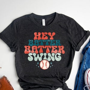 May include: A dark grey heather t-shirt with the text "Hey Batter Batter Swing" in a retro font. A baseball graphic is below the text.