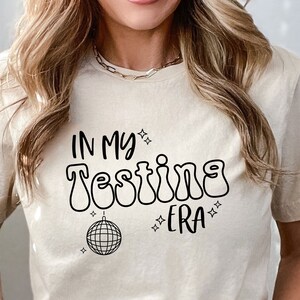 May include: A beige t-shirt with the text "In My Testing Era" in black with a disco ball graphic.