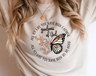May include: A beige sweatshirt with a circular design featuring a butterfly with pink, orange, and green wings. The design also includes flowers and the text "All my life you have been faithful, goodness of God, All my life you have been so good."