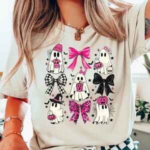 Puede incluir: Camiseta beige con un dise&ntilde;o tem&aacute;tico de Halloween. El gr&aacute;fico incluye varios personajes fantasma con acentos rosas, lazos y calabazas. El dise&ntilde;o tambi&eacute;n incorpora lazos a cuadros en blanco y negro, telara&ntilde;as y estrellas, creando una est&eacute;tica festiva y juguetona.