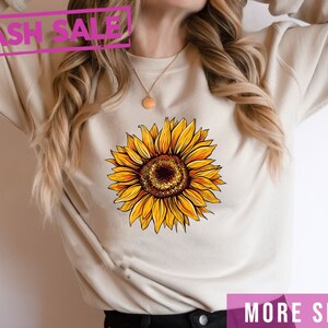 May include: A beige sweatshirt with a yellow sunflower graphic. The text "FLASH SALE" is in pink at the top of the image. The text "MORE SHIRT COLORS" is in pink at the bottom of the image.