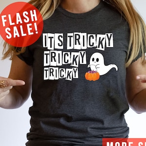 May include: Dark gray t-shirt with the text "IT'S TRICKY TRICKY TRICKY" in white block letters. A cartoon ghost and pumpkin graphic are on the shirt. A red circle with "FLASH SALE!" is in the upper left corner. The words "MORE SHIRT COLORS" are in a red rectangle.