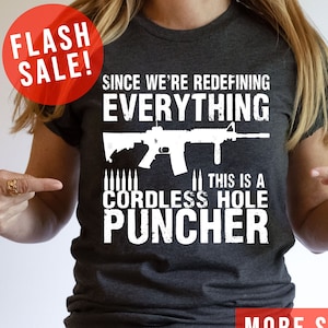 May include: A dark grey t-shirt with a white graphic of a rifle and the text "Since we're redefining everything... This is a cordless hole puncher".