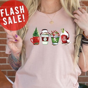 May include: A light pink t-shirt featuring four festive coffee cup designs with holiday themes. The cups are red, green, and white, with text that reads "Have a little holiday cheer" and "Dear Santa, I can explain". A red banner says "MORE SHIRT COLORS".