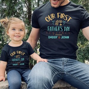 K&ouml;nnte beinhalten: Ein schwarzes T-Shirt mit dem Text "Our First Father's Day Together Daddy & John" in Gelb, Blau und Braun. Ein Kind tr&auml;gt eine kleinere Version des gleichen Shirts.