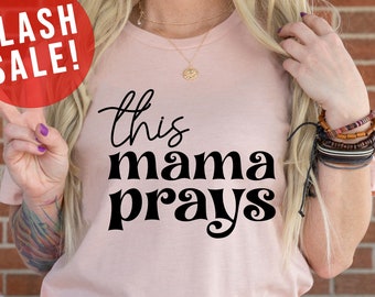 May include: A pink t-shirt with the text "this mama prays" in black lettering. The shirt is being worn by a person with a tattoo on their arm. The text is in a retro font style.