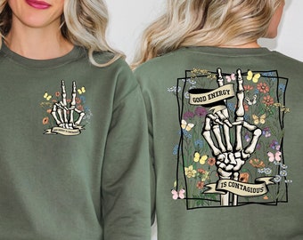 May include: A green sweatshirt with a skeleton hand making a peace sign surrounded by flowers and butterflies. The text "Good Energy is Contagious" is printed on a banner held by the skeleton hand.