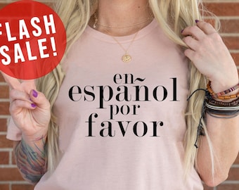 May include: A pink t-shirt with the text "en~ espanol por favor" in black lettering. The shirt is being modeled by a person wearing blue jeans.