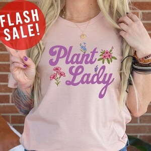 May include: A pink t-shirt with the text "Plant Lady" in purple script with floral accents. The shirt is being modeled by a person wearing blue jeans.